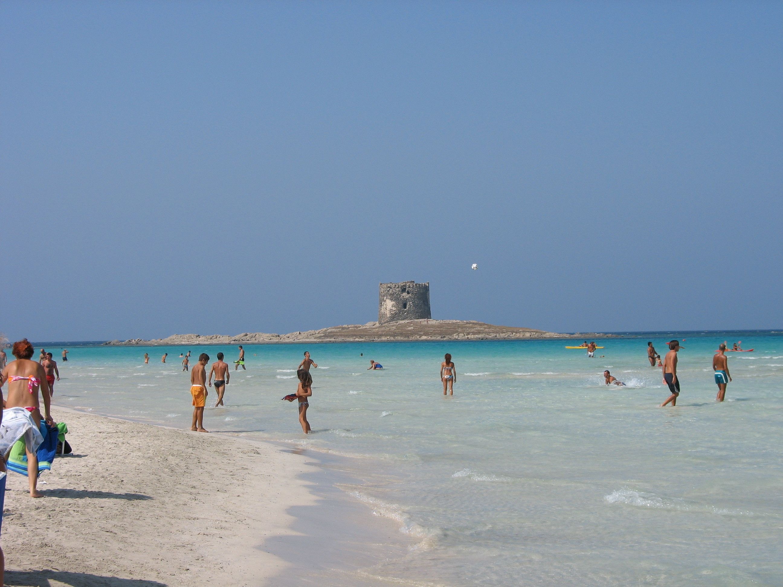 A beach with clear turquoise waters and a historic stone tower in the distance.