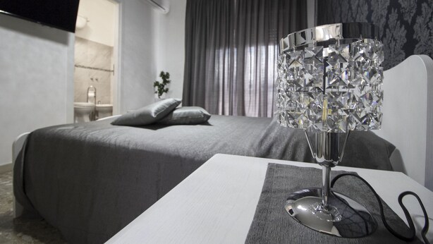 Habitación de hotel con una cama, una mesita de noche con lámpara, un televisor montado en la pared y un baño visible a través de una puerta abierta.