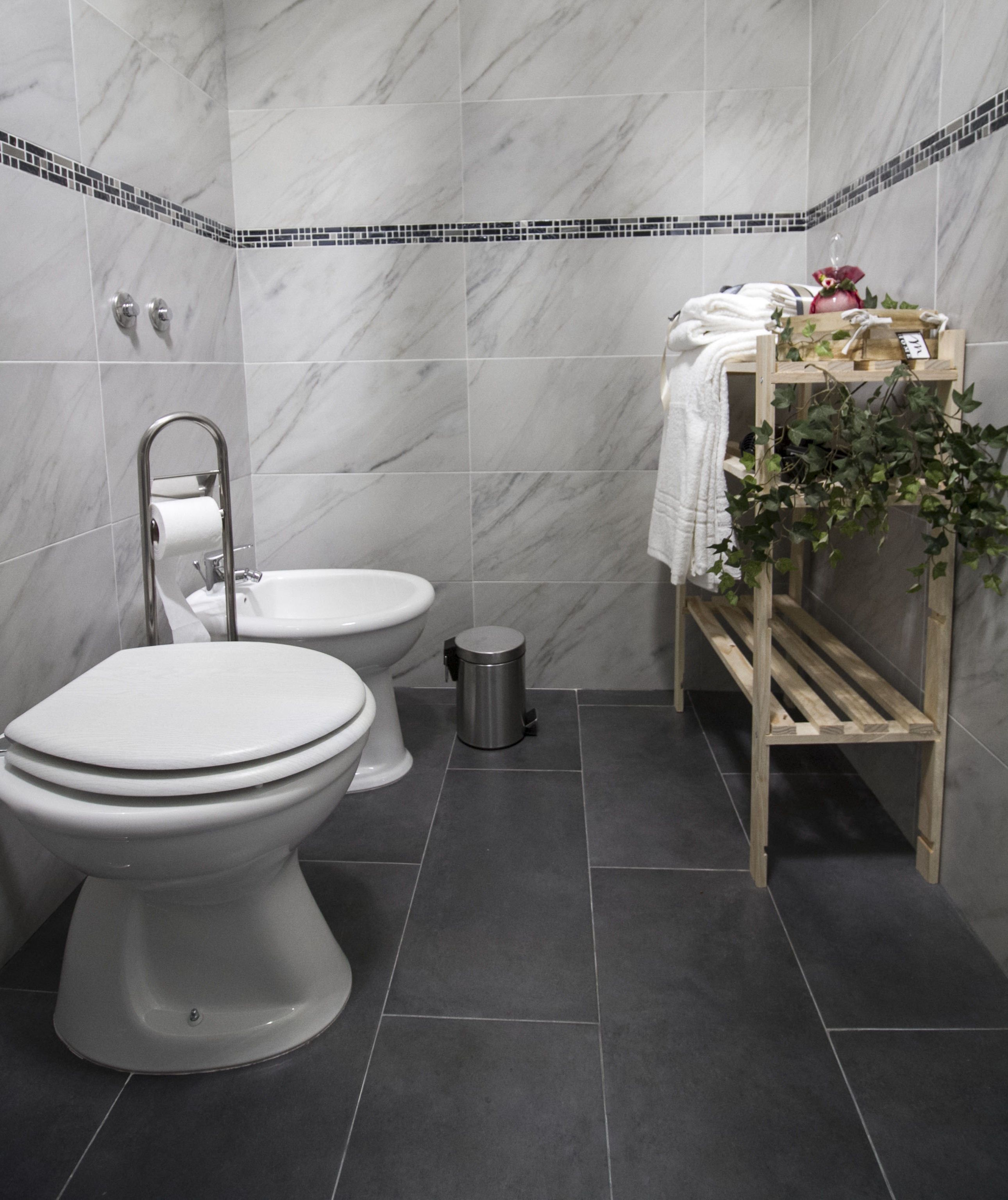 Un baño con dos inodoros, un lavabo, un cubo de basura y una repisa con toallas y una planta.