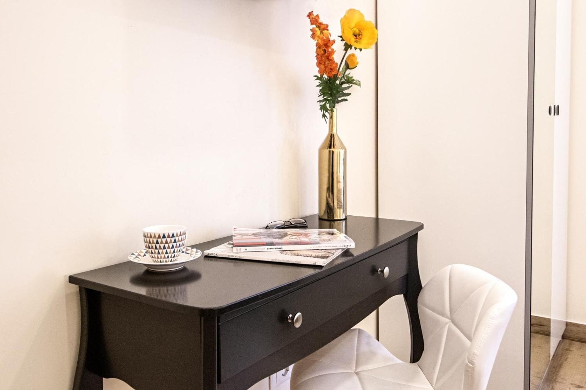 Un escritorio de madera oscura con una silla blanca, un florero con flores, una taza y algunas revistas.