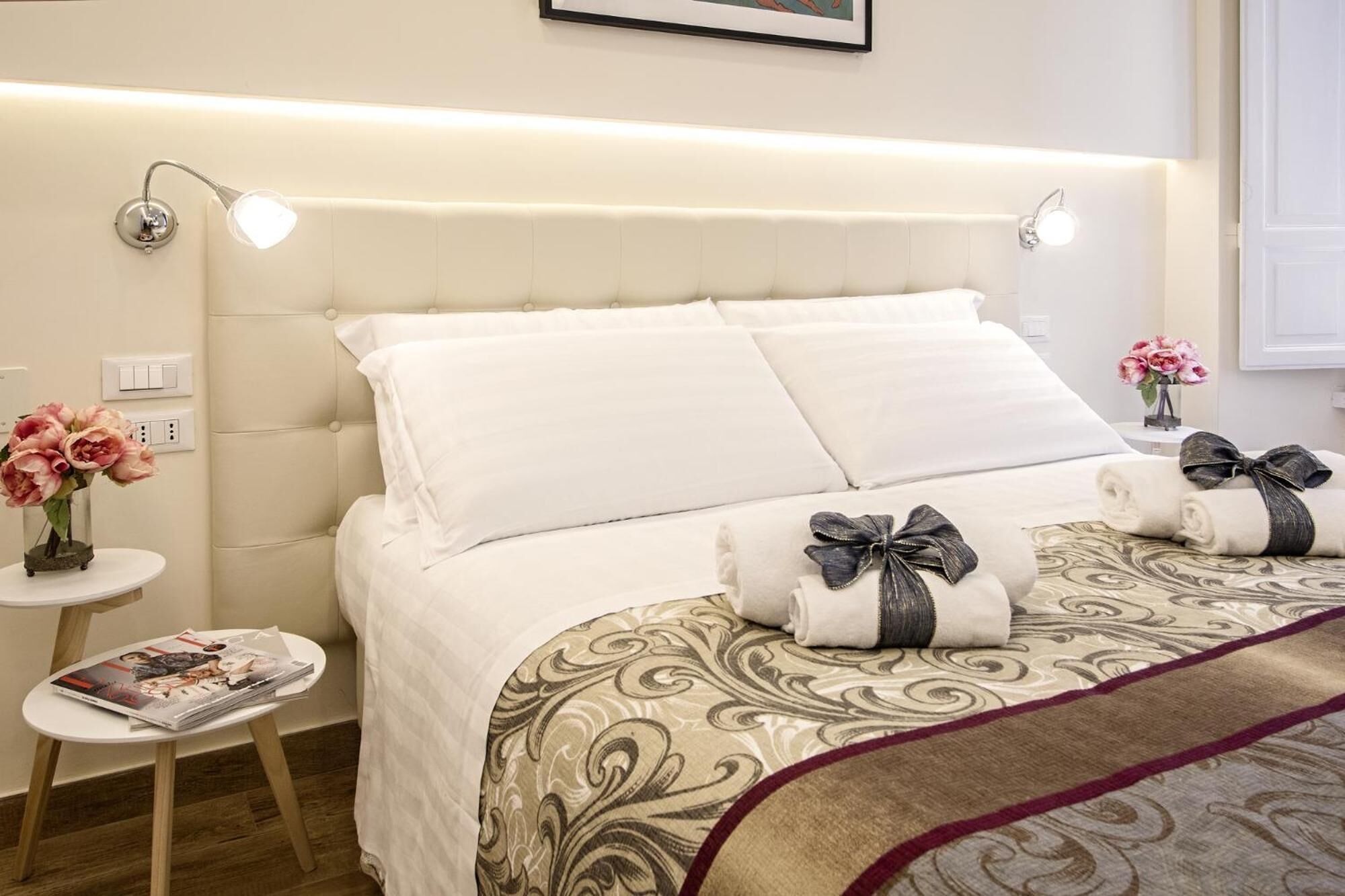 A neatly made bed with white linens and decorative pillows, a bedside table with a magazine, and a small vase with flowers.