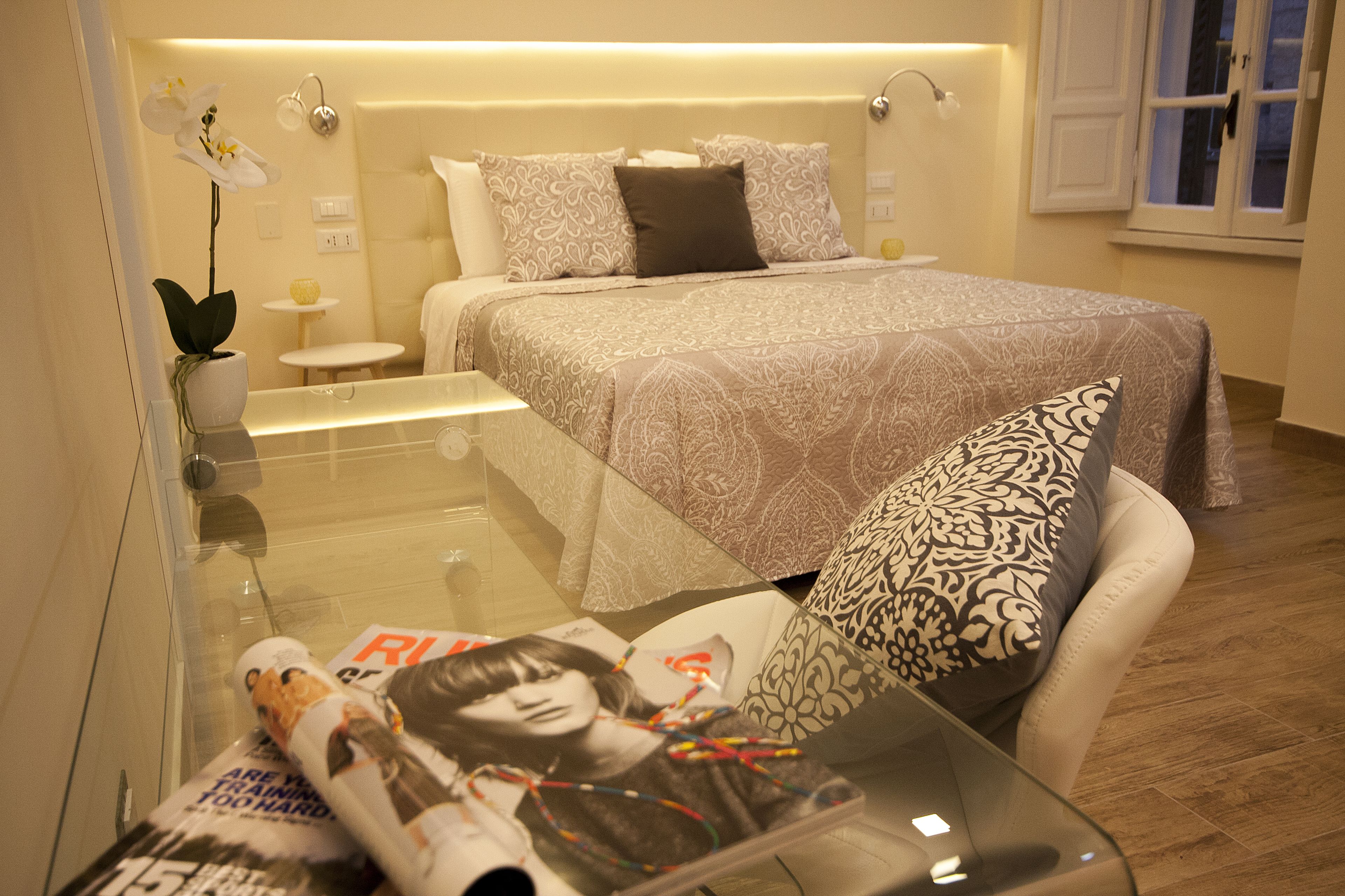 A modern bedroom with a glass table, a bed with patterned bedding, and a potted orchid.