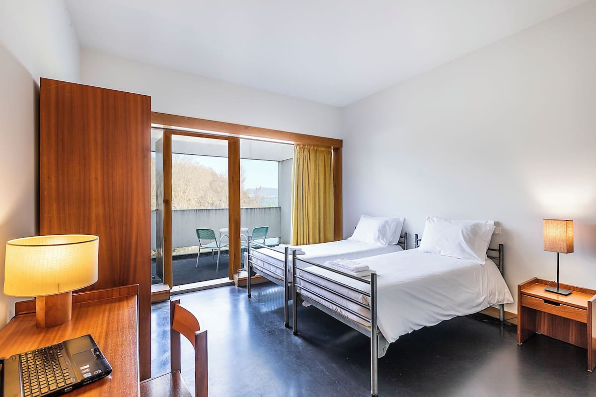 A hotel room with a bed, a desk with a laptop, a chair, a bedside table with a lamp, and a view of the outdoors.