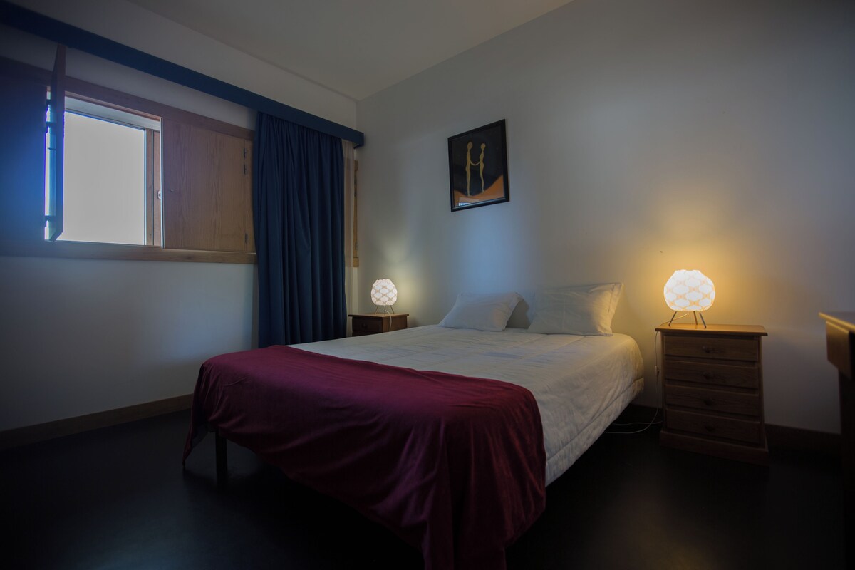 Un dormitorio con cama, dos mesitas de noche, un ventanal con cortinas, una obra de arte en la pared y una mesita de noche con lámpara.