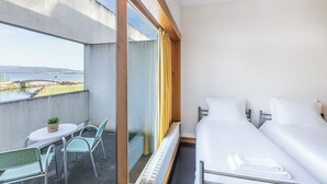 A balcony with a table and chairs, a bed with white linens, and a view of a bridge and water. - HI Viana Castelo - Pousada de Juventude - Hostel (Viana do Castelo)