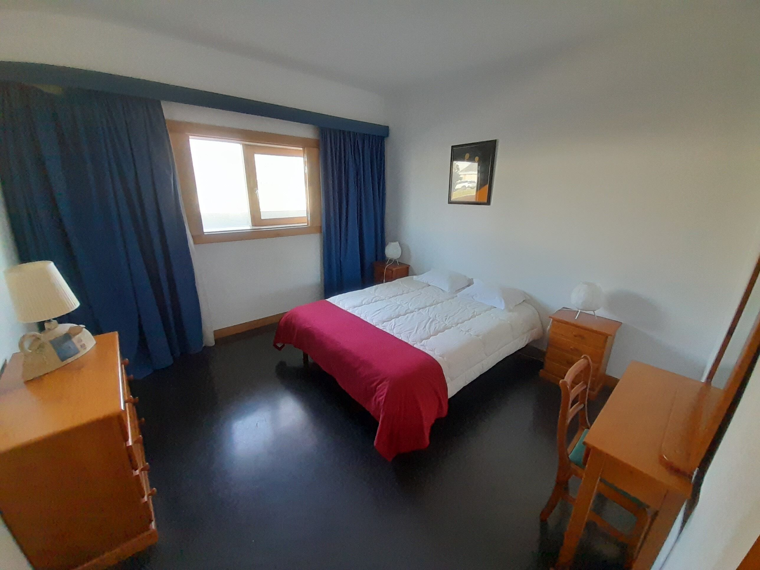 A bedroom with a bed, a desk, a chair, a nightstand, a lamp, a window with curtains, and a picture on the wall.