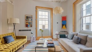 A living room with a yellow sofa, a white sofa, a striped rug, a small round ottoman, and a hanging light fixture.