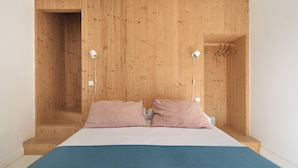 A bedroom with a wooden headboard, two bedside lamps, a bed with a blue bedspread, and a wooden wardrobe with a hanging rack.