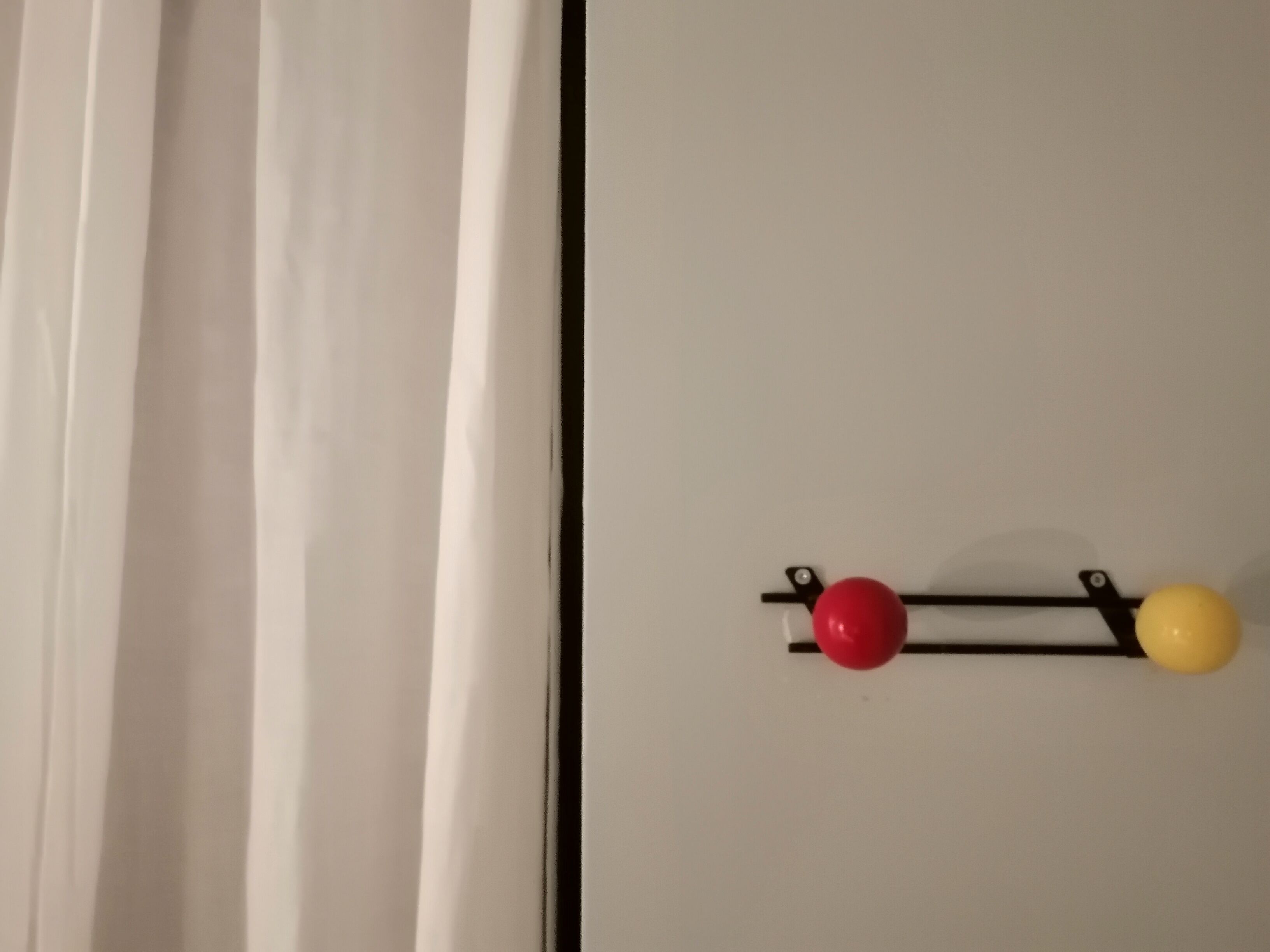 Una puerta blanca con marco negro y un pequeño gancho negro del que cuelgan una bola roja y una amarilla.