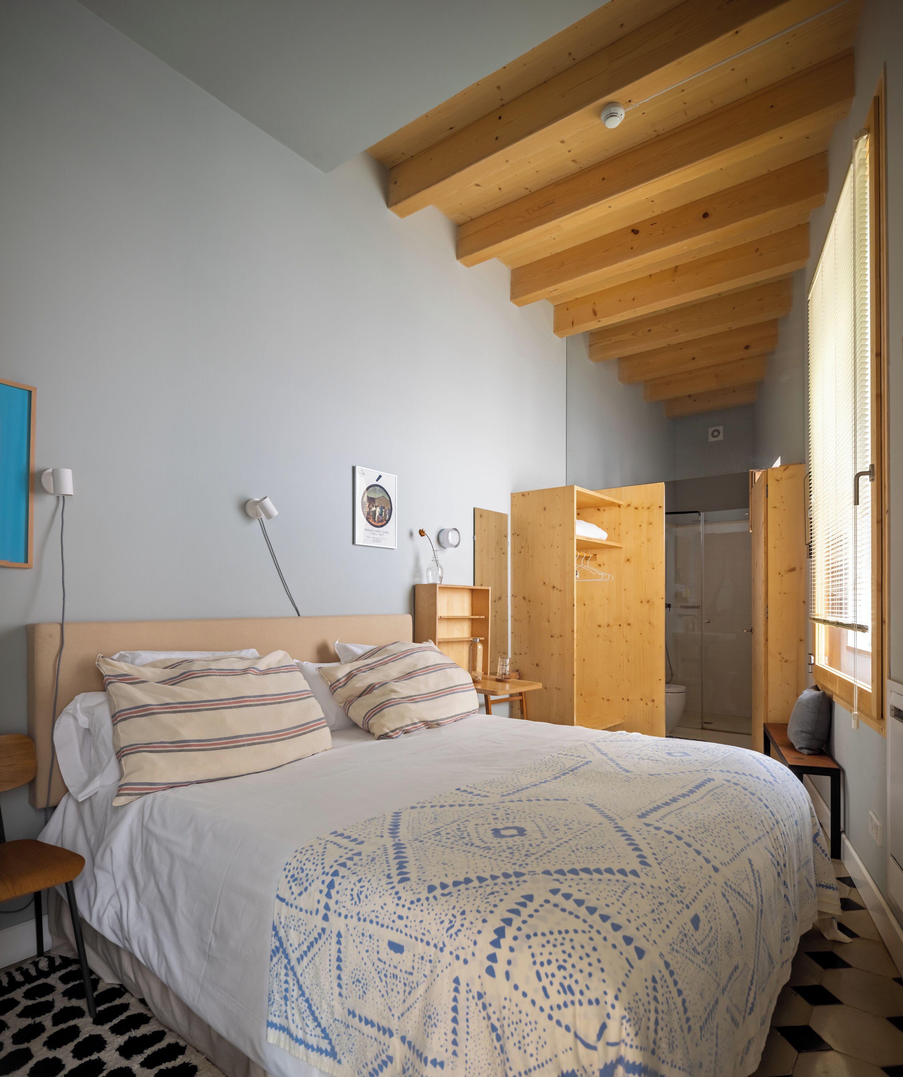 A bedroom with a wooden ceiling, a bed with striped pillows, a bedside table, a framed picture, and a window with blinds.
