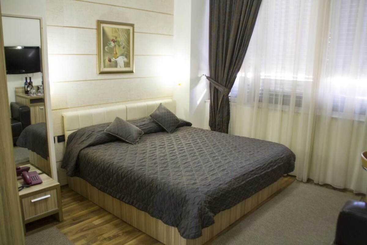 A bedroom with a bed, a bedside table, a mirror, a television, and a window with curtains.