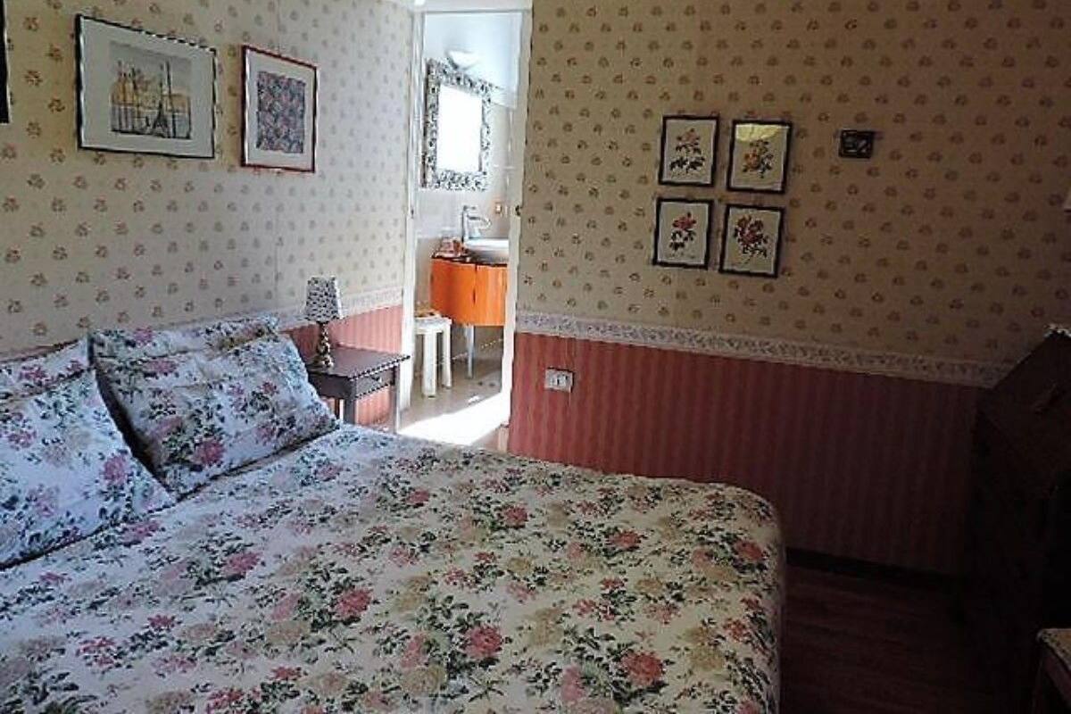 A bedroom with a floral bedspread, a small table, and a chair. There are framed pictures on the wall and a window with a view.