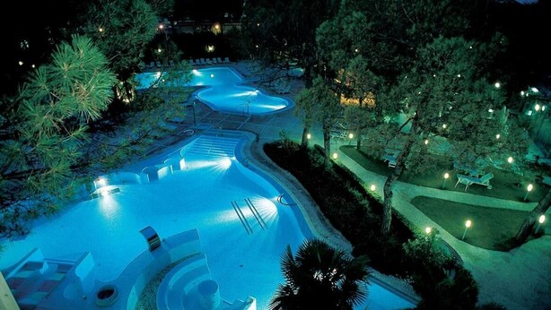 A large, winding swimming pool surrounded by trees and pathways, with lounge chairs and a small deck area. - B&B Dora e Flavio (Montegrotto Terme)