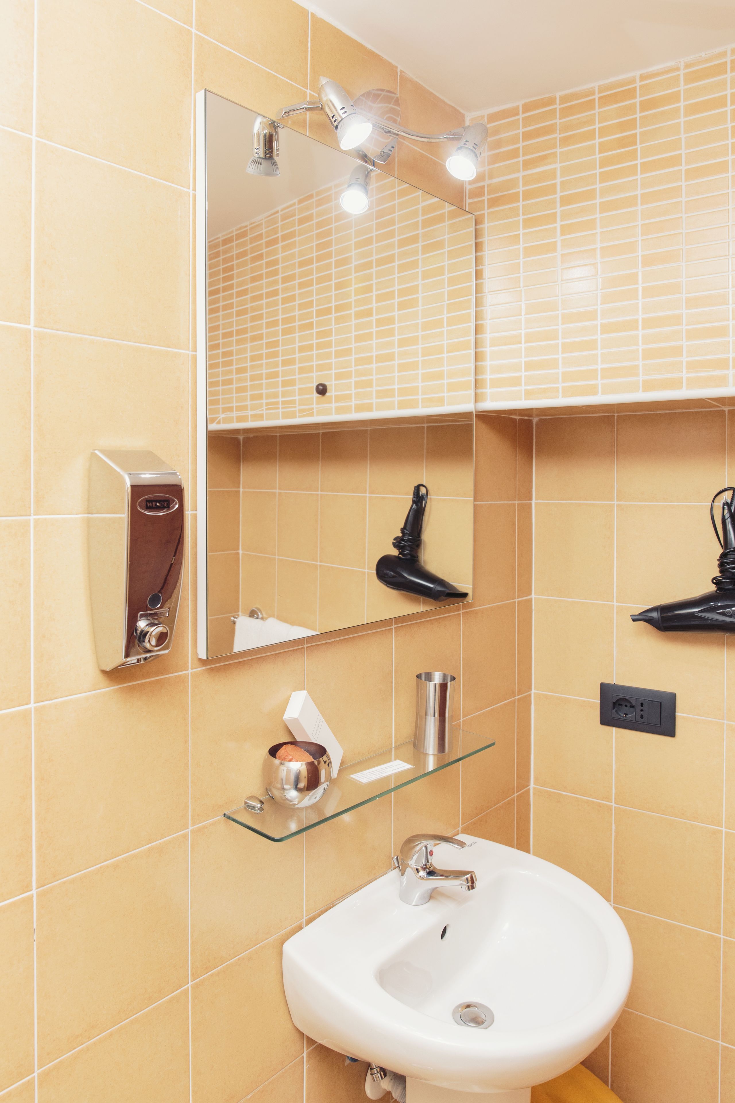 Une salle de bain avec un lavabo blanc, un miroir et une étagère en verre sur laquelle se trouve une bouteille.