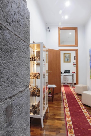 A hallway with a stone wall, a glass display case with items, a wooden door, a desk with a laptop, and a red carpet. - Palazzo Carafa della Spina - Charme (Naples)