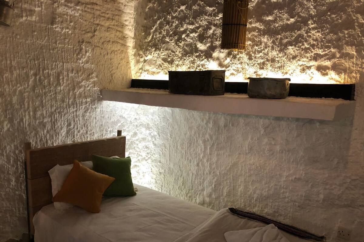 Una cama con cabecera de madera, ropa de cama blanca y dos almohadas en una habitación con paredes blancas texturizadas y una ventana con vista a árboles.