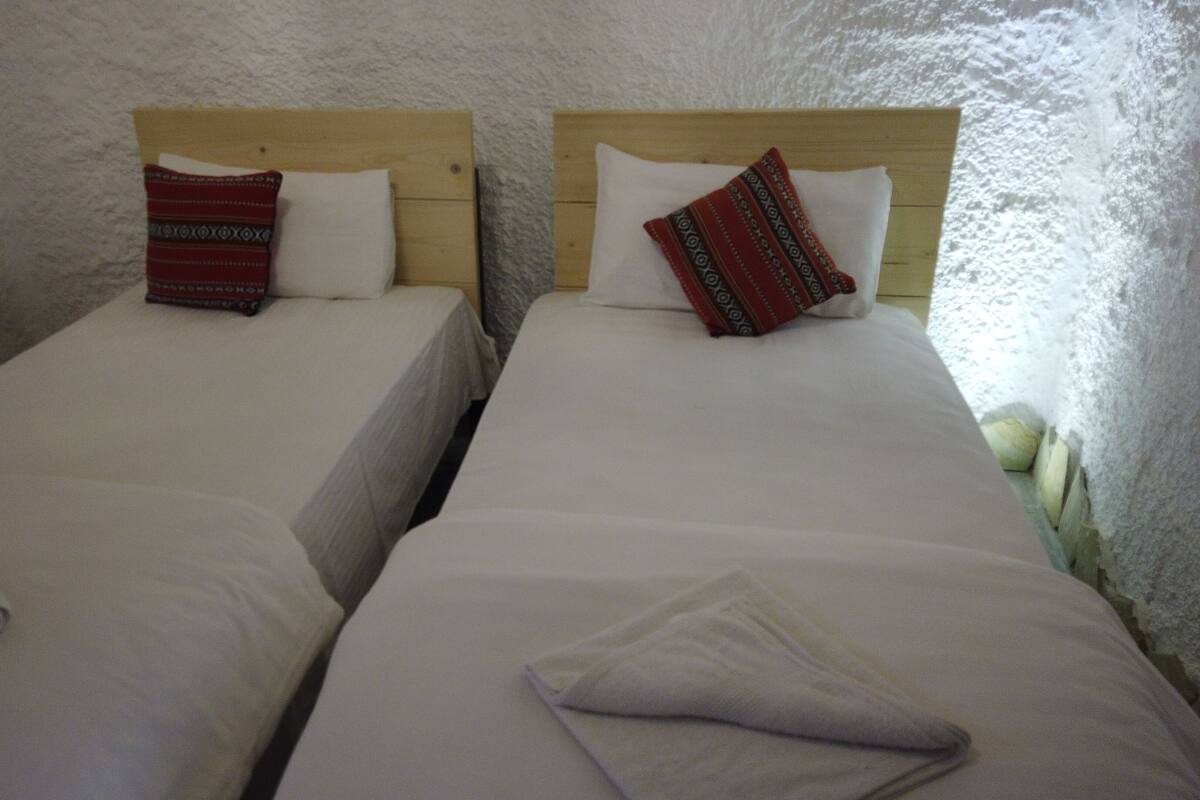 Dos camas individuales con cabeceros de madera y ropa de cama blanca, frente a una pared blanca con textura.