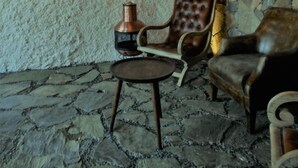 Un interior rústico con paredes de piedra, techo de madera y suelo de piedra. Se ve una mesa de madera con una silla, una pared de piedra con un pequeño ventanal y una lámpara colgante.