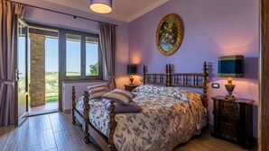 A bedroom with a large bed, a view of the outdoors, a painting on the wall, and a nightstand with a lamp.