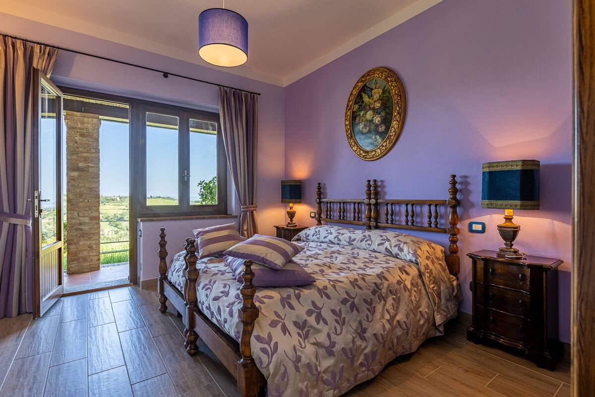Une chambre avec un grand lit, une vue sur l’extérieur, un tableau au mur et une table de chevet avec une lampe.