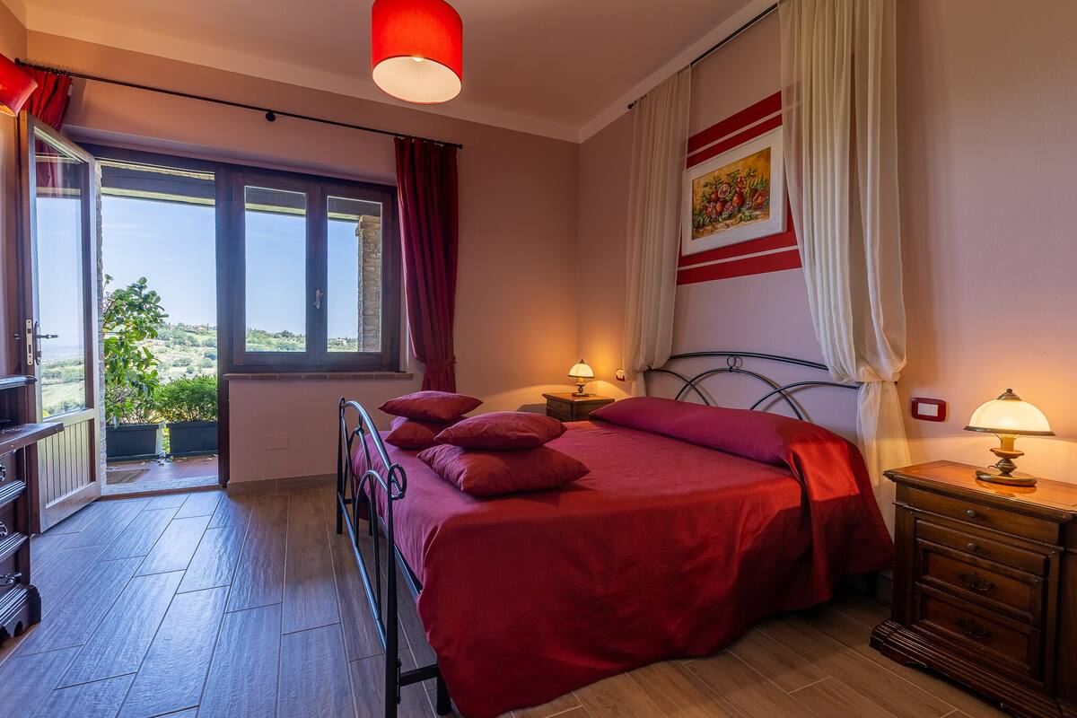 Une chambre avec un grand lit, une table de chevet avec une lampe, une fenêtre avec des rideaux et une vue sur la verdure à l’extérieur.