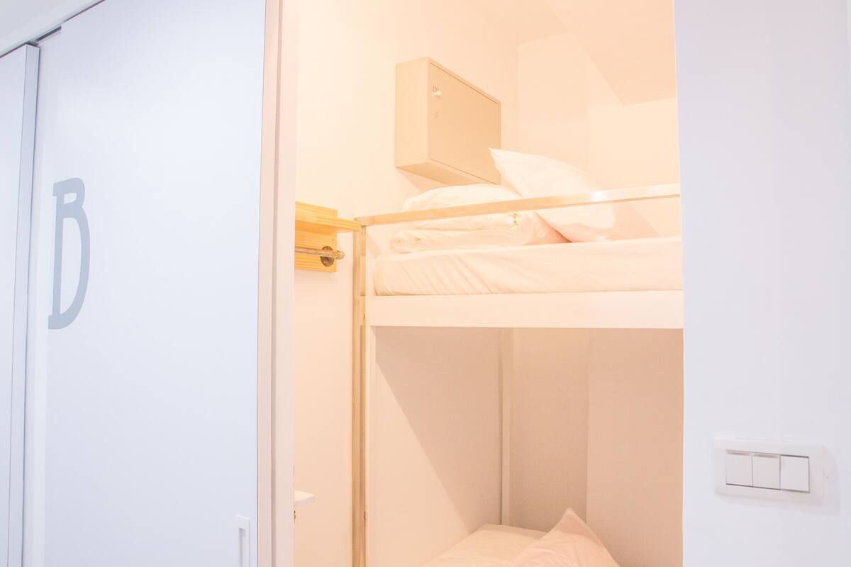 Un lit superposé avec une structure en bois et une literie blanche, un petit meuble en bois placé au-dessus, et un mur blanc avec un interrupteur.