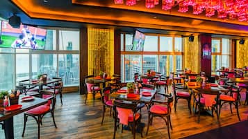 Um restaurante com várias mesas e cadeiras, janelas amplas com vista para a cidade e iluminação decorativa.