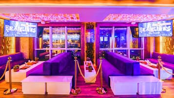 A luxurious bar area with purple seating, a minibar, and a view of the cityscape through large windows.