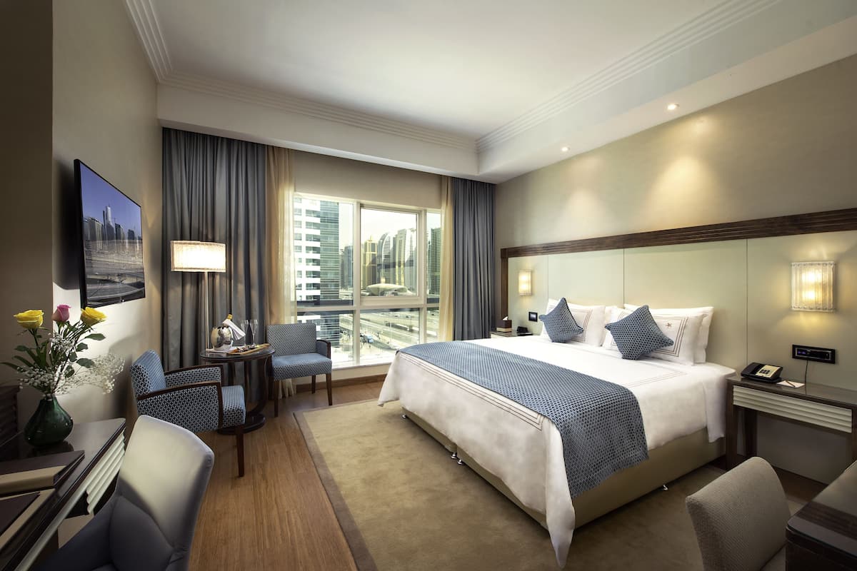 Une chambre d’hôtel avec un grand lit, un bureau, une chaise, une petite table avec une lampe, un vase de fleurs et une vue sur la ville par la fenêtre.