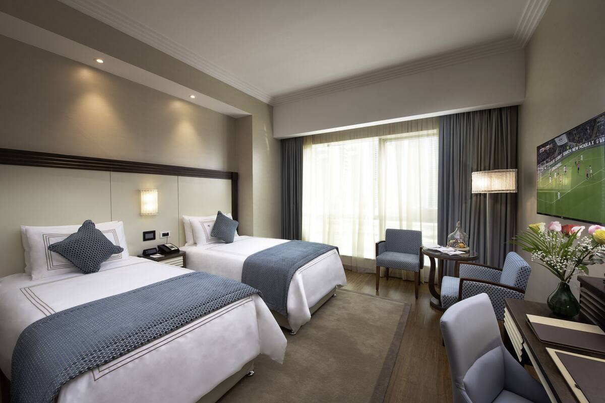 Une chambre d’hôtel avec deux lits, un bureau, une chaise et une grande fenêtre avec des rideaux.