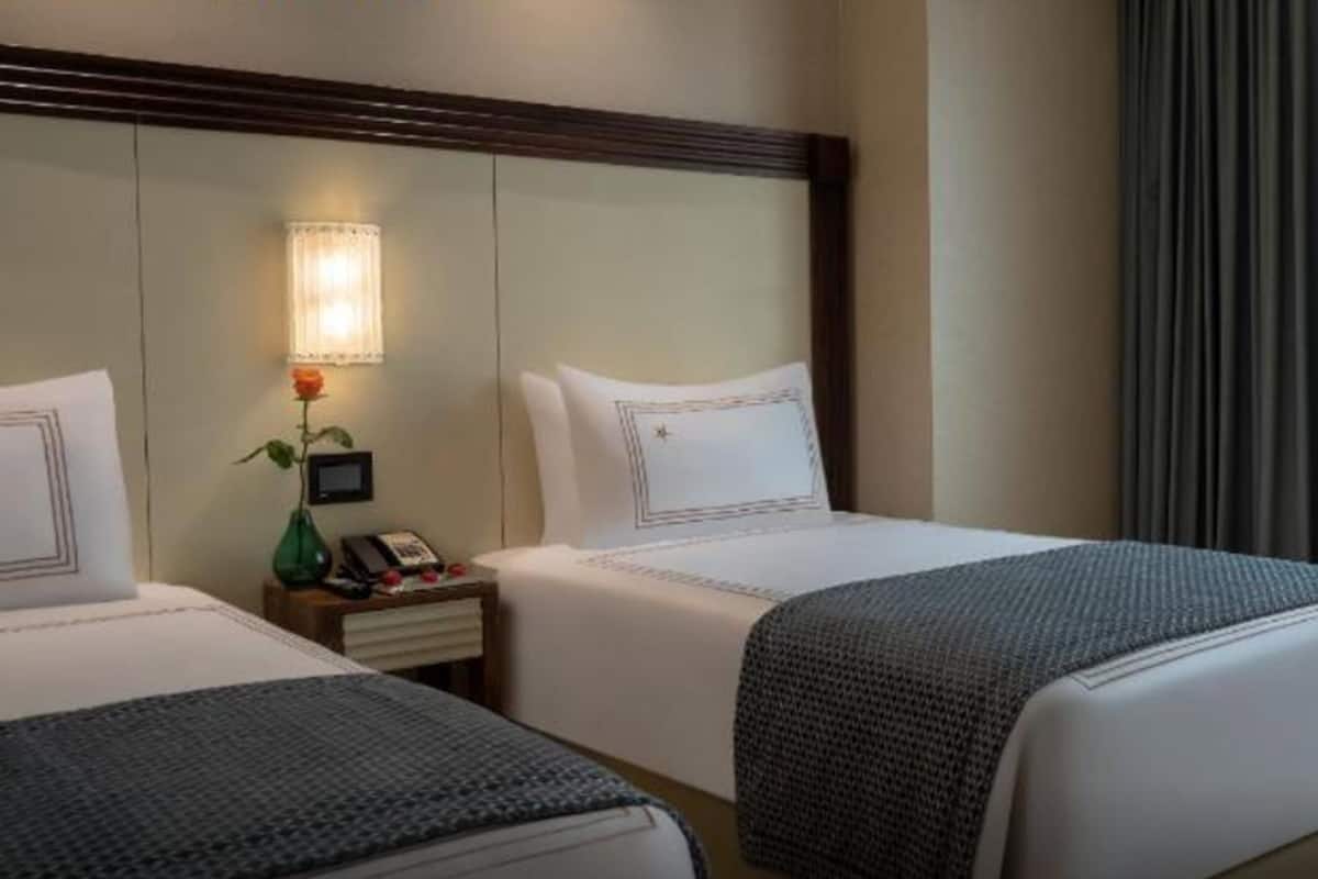 Une chambre d’hôtel avec deux lits, une table de chevet avec un téléphone et un vase contenant une fleur, une lampe, et une vue sur l’extérieur à travers des rideaux transparents.