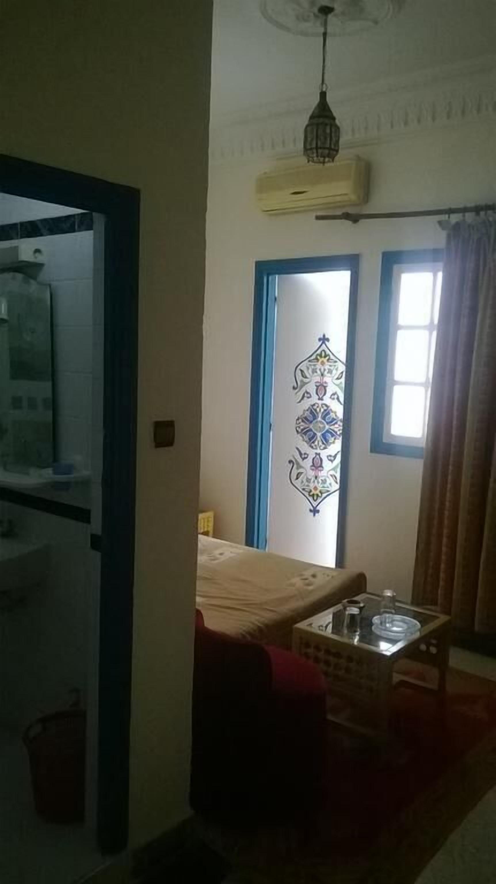 Ein Zimmer mit einem Bett, einem kleinen Tisch mit einem Glasbehälter, einem Fenster mit Vorhängen und einer dekorativen Wandverkleidung.