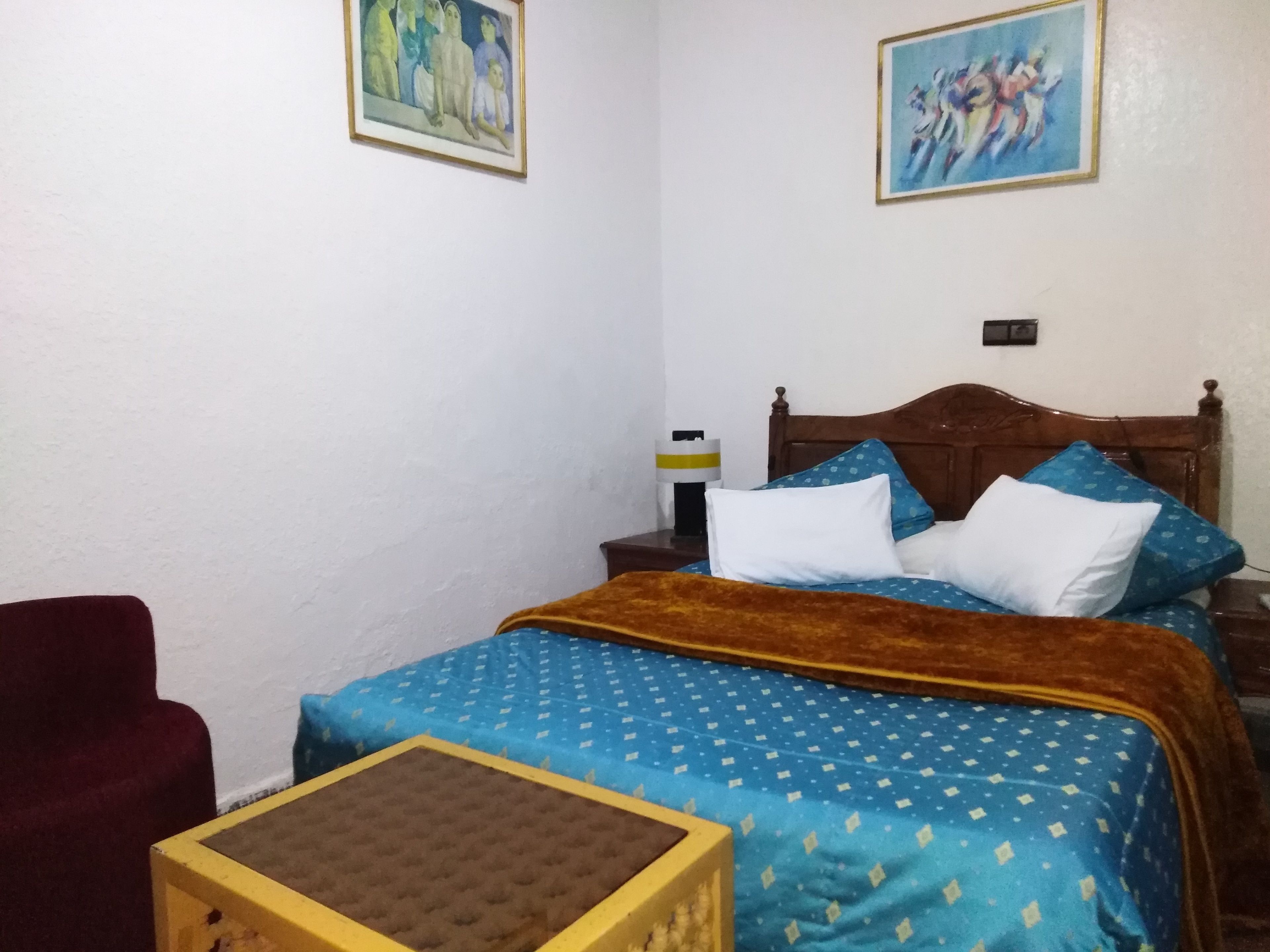 Un dormitorio con cama, cabecero de madera, mesita de noche, una silla roja y dos cuadros en la pared.