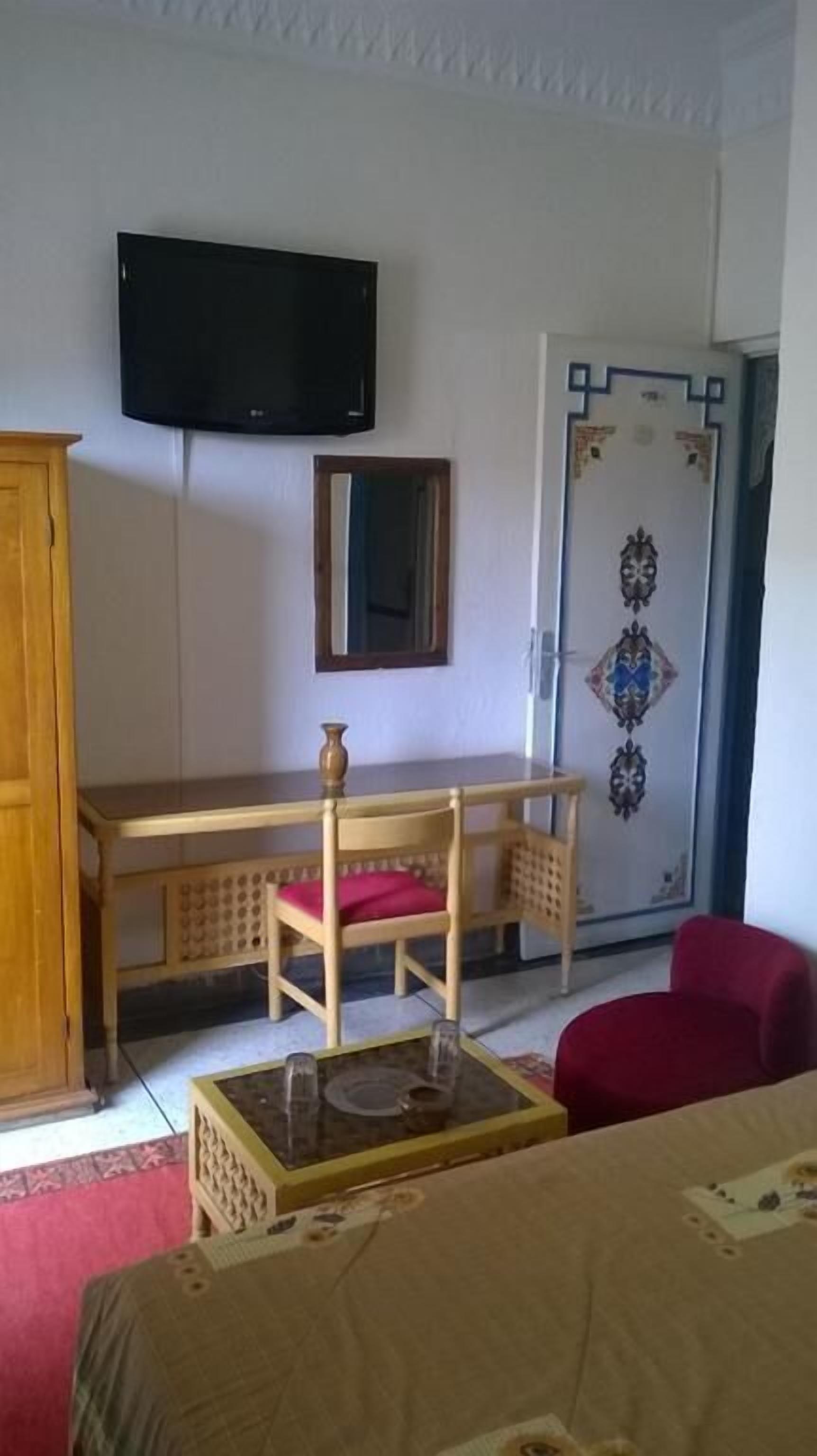 Una habitación con un escritorio de madera, una silla, un televisor montado en la pared, una puerta con diseños decorativos y una cama con cubrecama rojo.