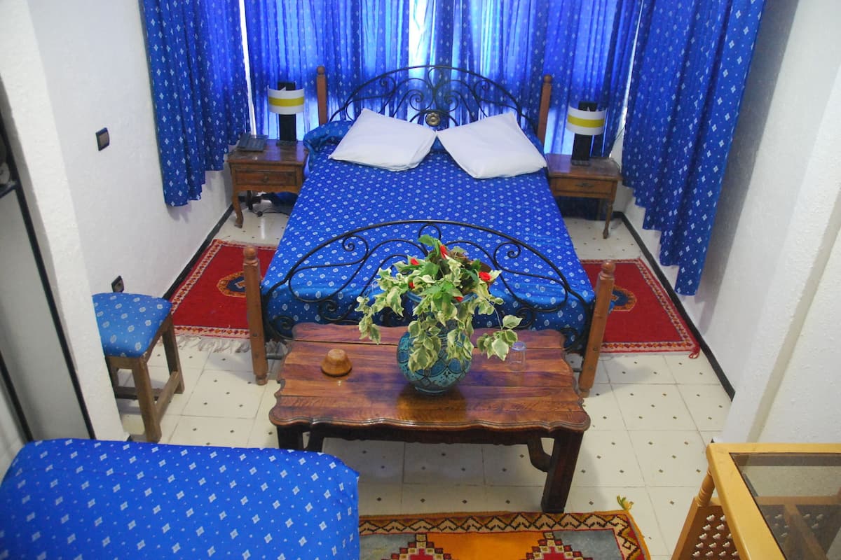 A bedroom with a bed, two bedside tables, a wooden nightstand, a chair, a table with a vase, and a patterned rug.