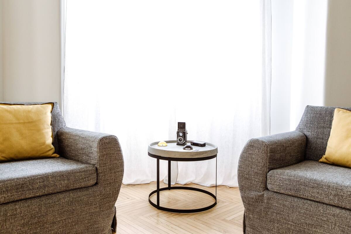 Ein modernes Wohnzimmer mit zwei grauen Sesseln, einem kleinen runden Couchtisch mit einem Vintage-Radio und einem weißen Vorhang im Hintergrund.