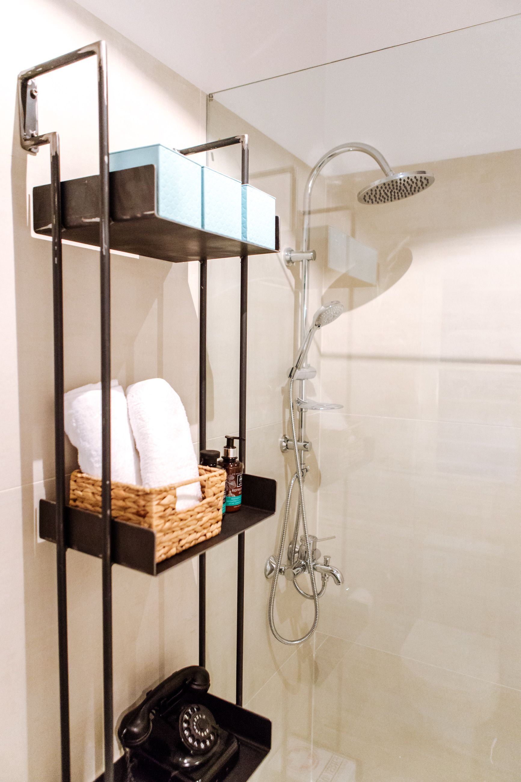 A modern bathroom with a glass-enclosed shower, a black telephone, and a shelf with towels and toiletries.