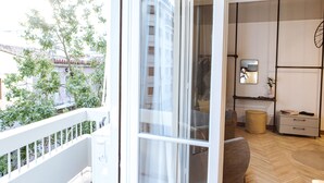 A balcony with a white railing, a view of trees and buildings, and a room with a wooden floor, a sofa, a small table, and a cabinet. - We Saw Athens (Athens)