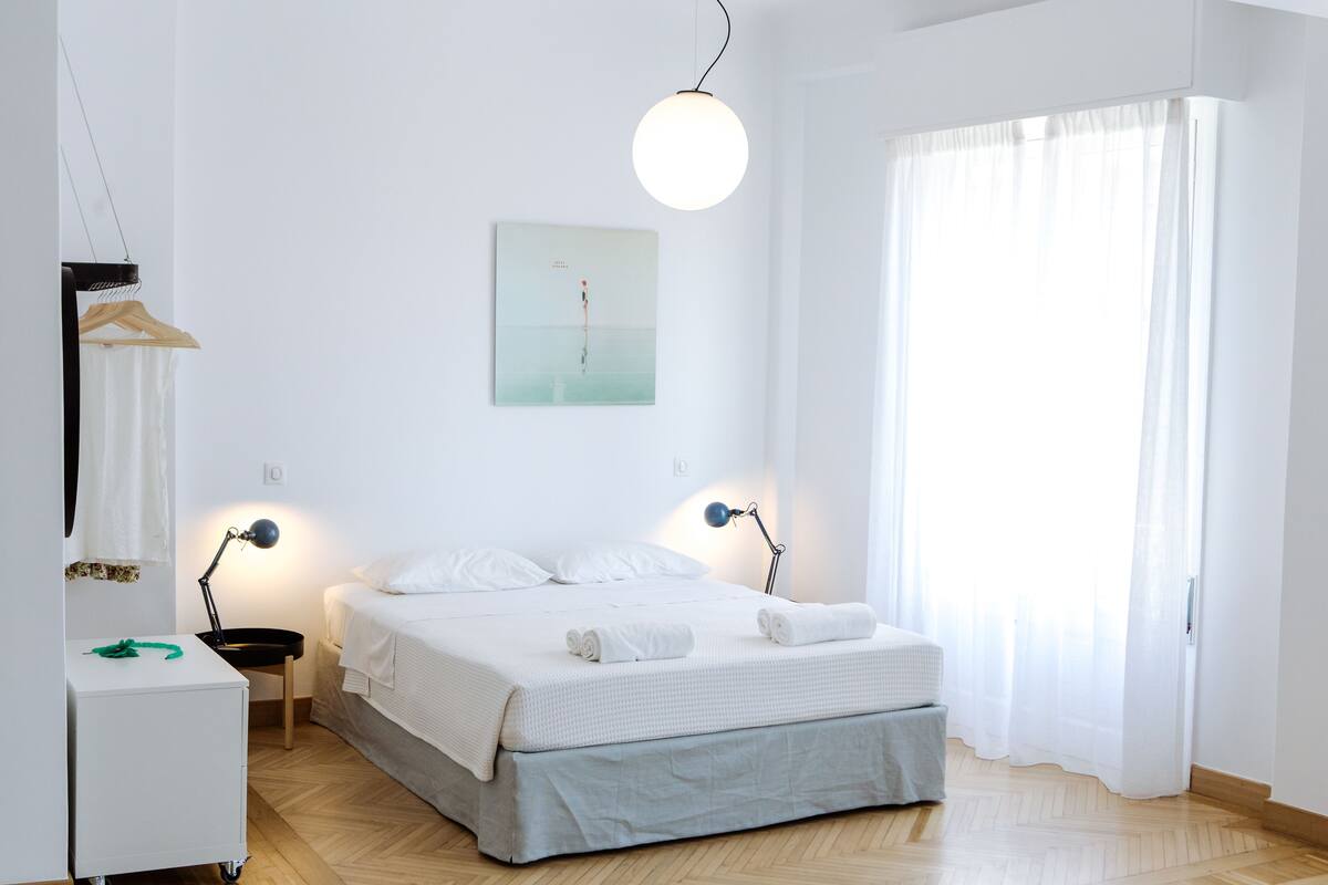 Ein schlicht eingerichtetes Schlafzimmer mit Bett, Nachttischlampen, einer Pendelleuchte, einem Gemälde und einem Fenster mit Vorhängen.