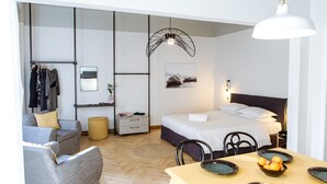 A modern hotel room with a bed, a dining table with plates, a chair, a sofa, a mirror, a coat rack, and a hanging light fixture. - We Saw Athens (Athens)