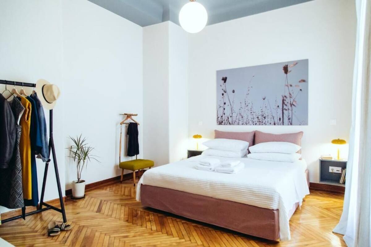 Ein Schlafzimmer mit Bett, Nachttischen, einem Kleiderschrank, einer Kleiderstange, einer Topfpflanze und einem Bild an der Wand.