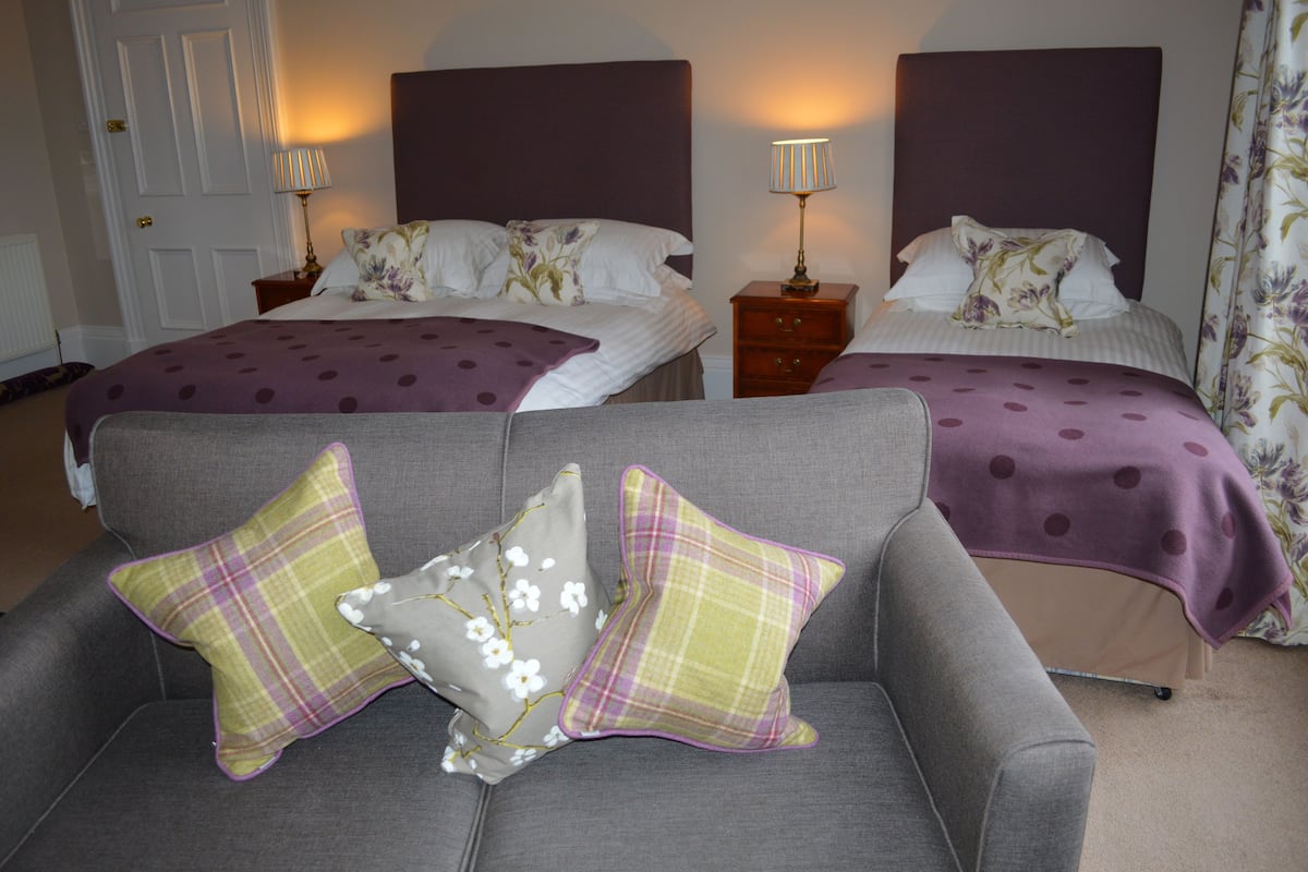 A hotel room with two beds, each with a headboard, bedside tables, and lamps. There is a grey sofa with two decorative pillows in front of the beds.