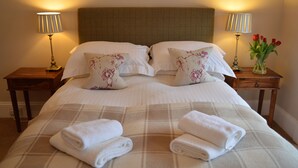 Cama bem arrumada com duas almofadas, duas mesas de cabeceira com abajures e um vaso com flores vermelhas no lado direito.