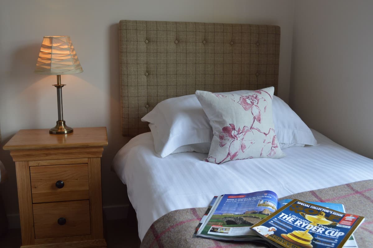 A neatly made bed with white linens and a floral pillow, a bedside table with a lamp, and a magazine on the bed.