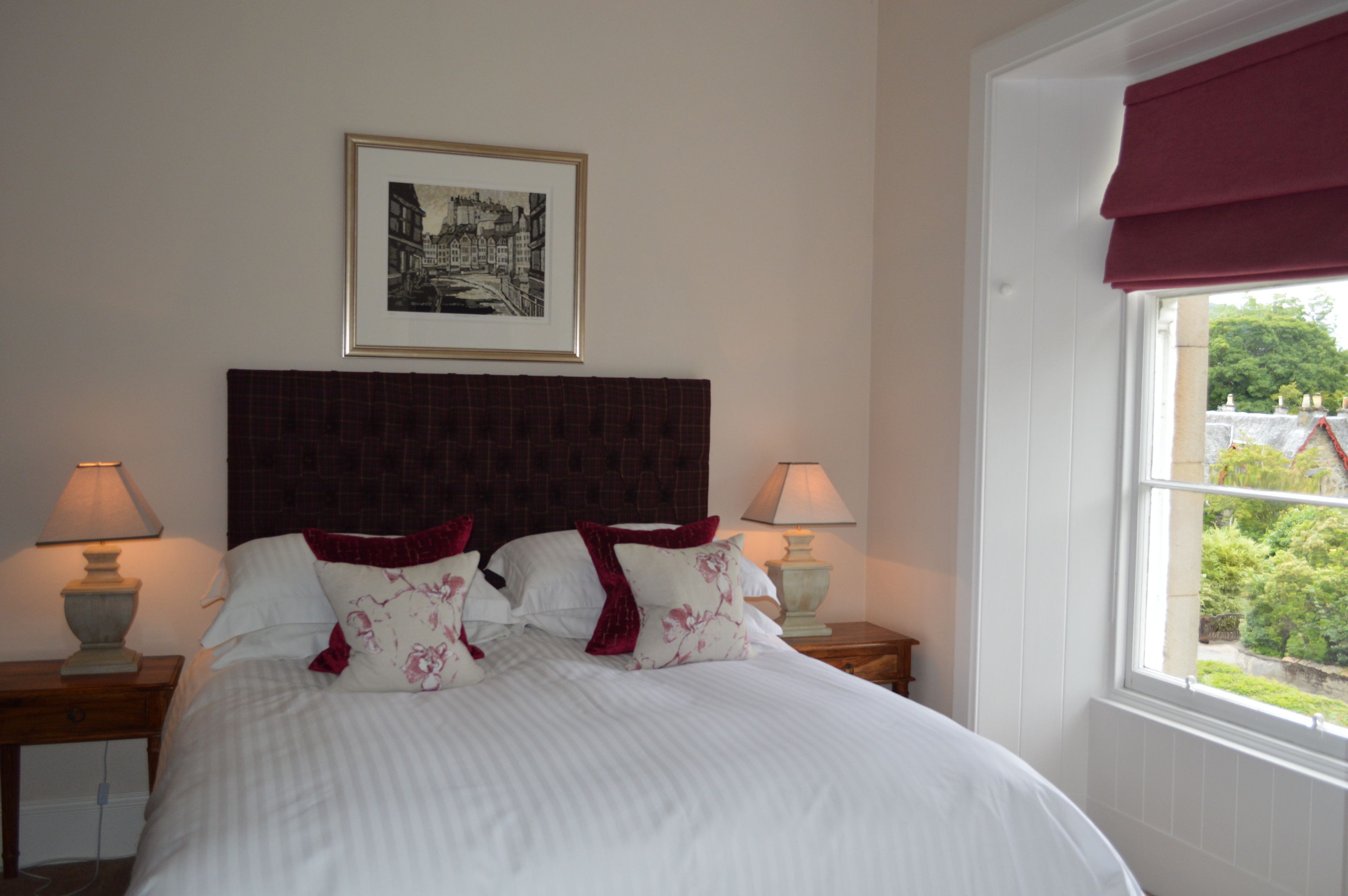 A neatly made bed with white linens and floral pillows, a framed black and white cityscape picture on the wall, and a window with a red valance.