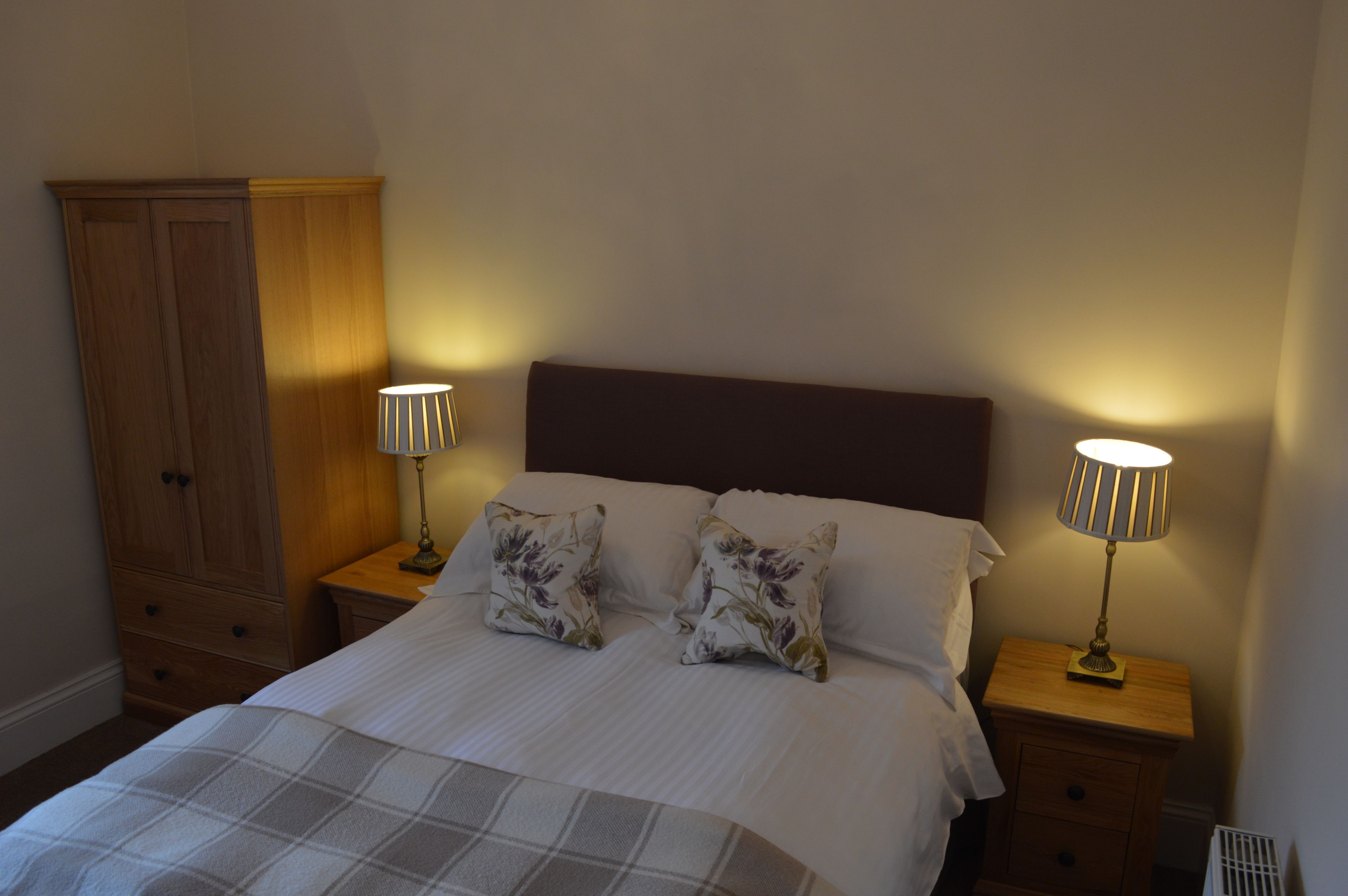 Una cama bien hecha con dos almohadas, un armario de madera y dos lámparas de noche sobre mesitas de madera.