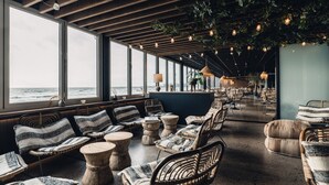 A spacious lounge area with wicker furniture, large windows overlooking the ocean, and hanging light fixtures. - Ocean Hotel (Falkenberg)