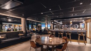 A modern bar area with a round wooden table and bar stools, a seating area with a sofa and chairs, and a view of the city through large windows.