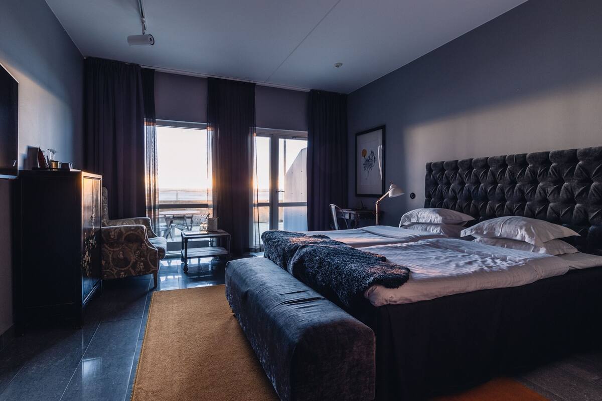 Una habitación de hotel moderna con una cama grande, una zona de estar con sofá y una mesita, un escritorio y vistas al exterior a través de amplios ventanales.