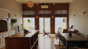 A spacious living area with a large wooden dining table, a kitchen with white cabinetry, and a view of the ocean through large sliding doors. - Ocean Hotel (Falkenberg)